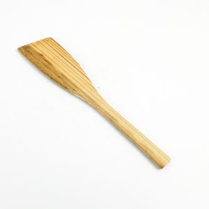 May include: A wooden spatula with a rectangular head and a long, rounded handle. The head features parallel grooves, and the spatula is made of light-colored wood. The spatula is angled diagonally against a white background.