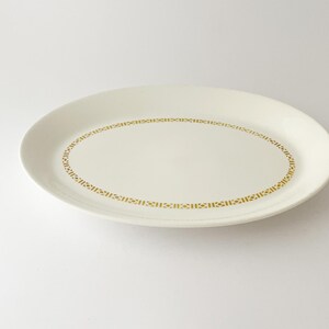 May include: An oval, off-white serving platter with a gold geometric border. The plate is made of ceramic and is set against a white background. The design is simple and elegant, suitable for various dining occasions.