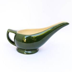 May include: A green ceramic gravy boat with a handle and a spout. The interior is a cream color, contrasting with the glossy exterior. The gravy boat sits on a small, round base.