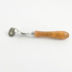 May include: A sewing tracing wheel with a wooden handle and a metal wheel with small, sharp teeth. The wheel is attached to a curved metal arm. The handle is a light brown color.