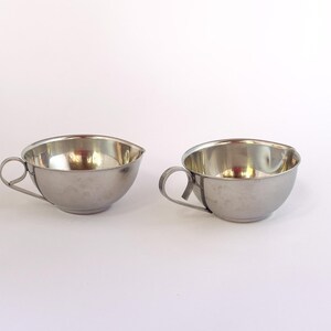 May include: Two stainless steel cups with handles. The cups have a rounded bowl shape and a shiny, reflective surface. The handles are a simple, curved design. The cups are likely for serving beverages.