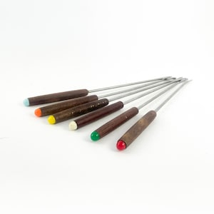 May include: Set of six fondue forks with dark brown wooden handles and stainless steel tines. Each handle has a different colored tip: blue, orange, yellow, white, green, and red. The forks are arranged diagonally on a white surface.