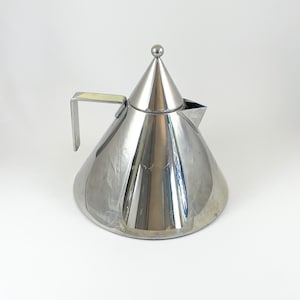 May include: A polished stainless steel teapot with a conical body and a matching conical lid topped with a small spherical finial. The teapot features a flat, angular handle extending from the side and a short, triangular spout for pouring.