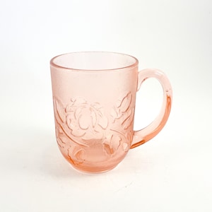May include: A pink glass mug with a textured surface and a floral design. The mug has a handle and is suitable for hot or cold beverages. The mug is a light pink color.