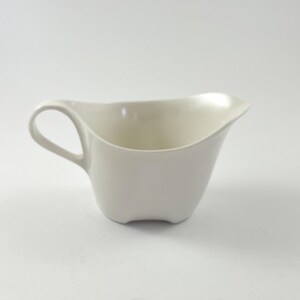 May include: A solid off-white ceramic gravy boat featuring a smooth, curved body with a single integrated handle on the left side and a wide, tapered pouring spout on the right. The base has a subtle indented design, and the interior is deep.