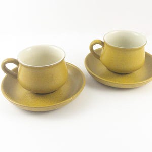 May include: Two matching mustard-yellow ceramic teacups with saucers. The cups have a rounded shape with a handle and a cream-colored interior. The saucers are circular and have a slightly raised edge. The set is on a white background.
