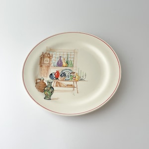 May include: A cream-colored decorative plate with a red rim. The plate features a watercolor illustration of a room with a grandfather clock, vases, and other decorative items. The plate is round.