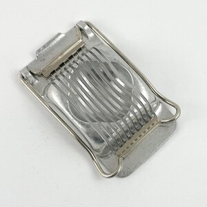 May include: A vintage, stainless steel egg slicer with a rectangular base and parallel wires for slicing. The slicer has a hinged top and a curved handle. The metal has a slightly worn appearance.