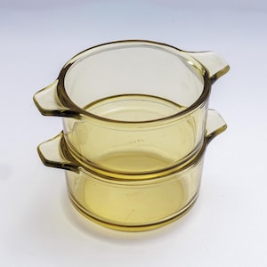 May include: Two amber-colored glass Pyrex bowls stacked on top of each other. Each bowl has a rounded shape with two handles. The bowls are made of transparent glass, allowing the contents to be seen.