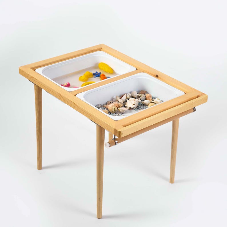 May include: A wooden sensory table with two white plastic bins. One bin contains colorful toys, and the other is filled with seashells and small rocks. The table has light-colored wooden legs and a frame.