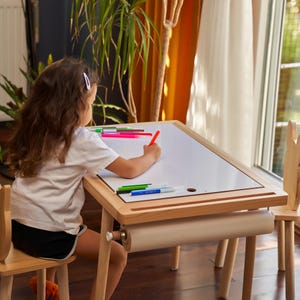 May include: A child is seated at a wooden art table with a white surface, drawing with a red marker. Several colorful markers are on the table. The table has a roll of paper underneath. A wooden chair is next to the table.
