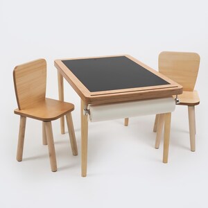 May include: A child's wooden table and two chairs set. The table has a black chalkboard surface and a roll of paper underneath. The chairs and table are made of light-colored wood. The set is designed for children's activities and play.