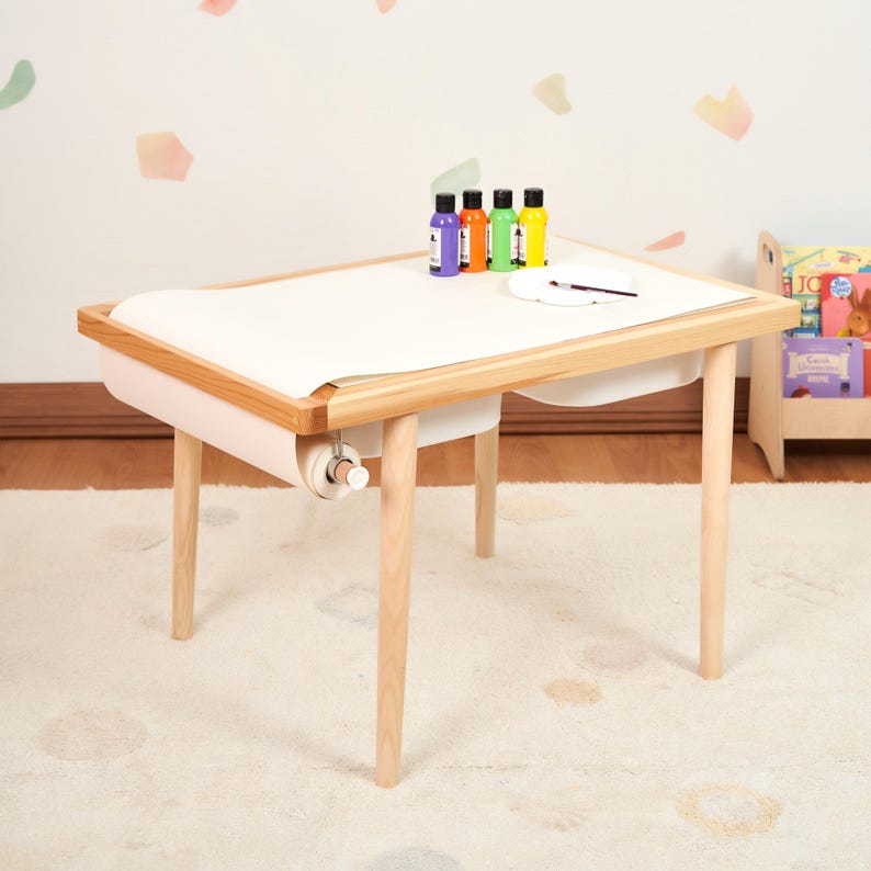 May include: A light wood art table with a white paper roll and a built-in tray. The table has four legs and a wooden frame. Several bottles of paint and a paintbrush are on the table. The table is on a light-colored rug.