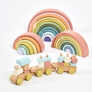 Op de afbeelding: Een houten speelgoedtrein met drie verbonden wagons voorzien van pastelkleurige blokken en wielen, vergezeld door drie in elkaar passende regenboogvormige stapelspeeltjes in zachte tinten roze, geel, groen, blauw en paars op een witte achtergrond.