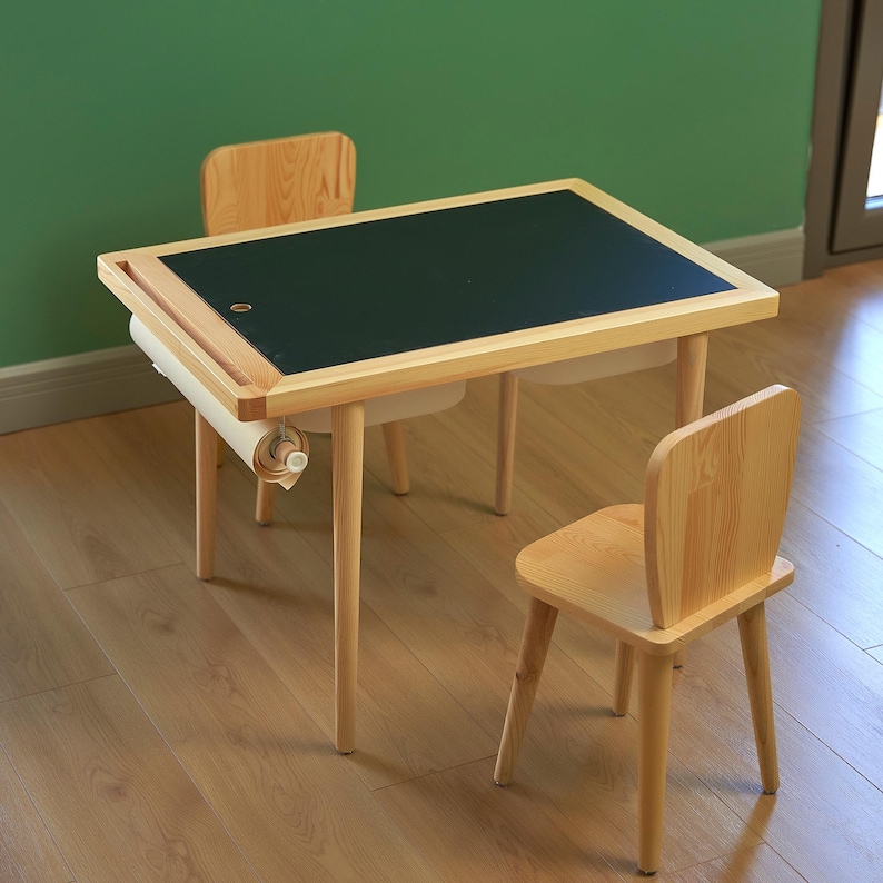 May include: A light wood children's table and two matching chairs. The table has a black chalkboard surface and a paper roll holder. The chairs have a simple, curved back design. The table is approximately 30 inches long.