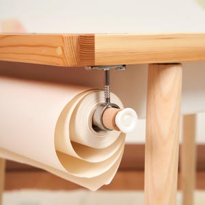 May include: A roll of off-white paper suspended under a light-colored wooden table. The paper is held by a metal and wood mechanism with a white knob. The table has a natural wood finish.