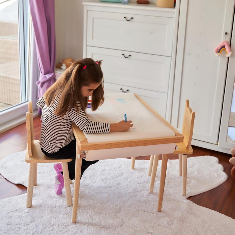 May include: A light wood children's table and chair set. The table has a white paper roll and a light pink trim. The chair has a cutout design at the top. The child is drawing with a blue marker on the paper.