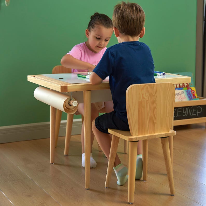 May include: A light wood children's table with a roll of paper attached, and two matching chairs. Two children are seated at the table, drawing with markers. The table has a white surface and a wooden frame.