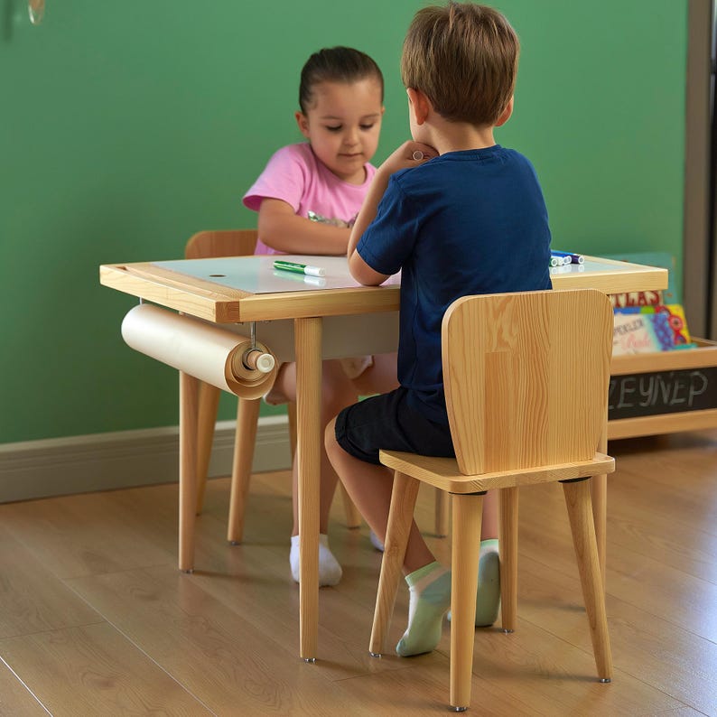 May include: A light wood children's table with a white top and a roll of paper attached. Two children are seated at the table, one wearing a pink shirt and the other a blue shirt. The table has a matching wooden chair.