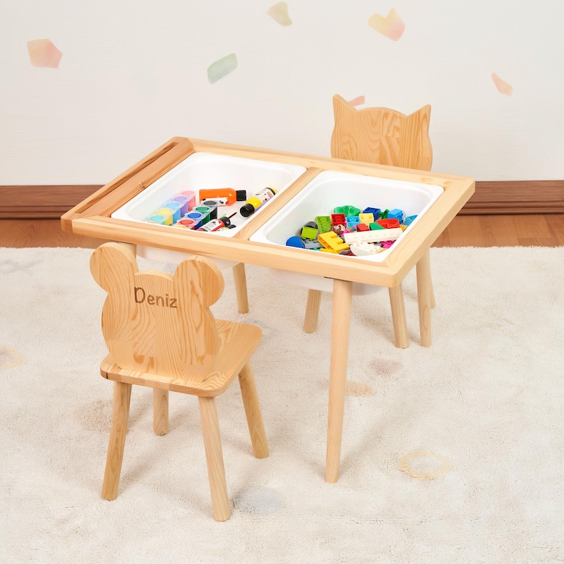 May include: A wooden children's play table with two white bins, filled with toys. The table has a natural wood finish and is paired with two matching chairs. One chair has a bear-shaped back and the name "Deniz" engraved on it.