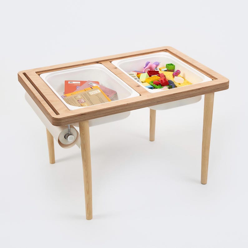 May include: A wooden sensory table with two white plastic bins. One bin contains colorful toys, the other has books. The table has light wood legs and a paper towel holder.