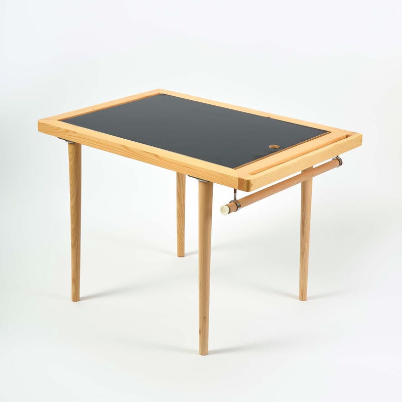 May include: A small wooden desk with a black top and light-colored wooden legs. The desk has a rectangular shape with a built-in tray and a wooden bar for hanging items. The desk is approximately 30 inches long and 20 inches wide.