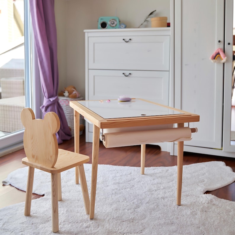 May include: A light wood children's table with a paper roll and a matching chair with a bear-shaped back. The table has a white surface and is set on a white rug. The chair and table are made of light-colored wood.