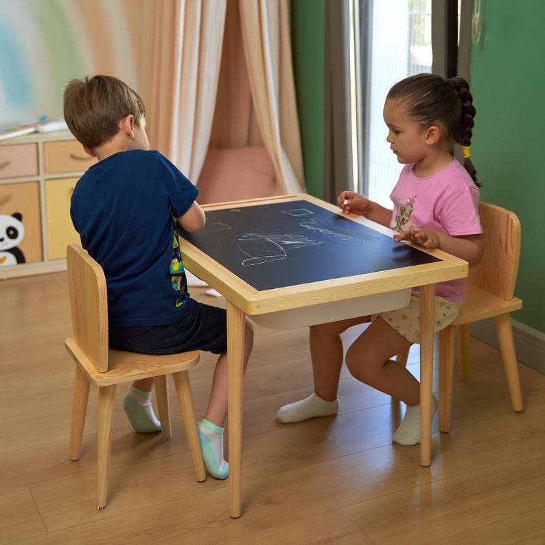 May include: A wooden children's table with a black chalkboard surface and a white storage bin underneath. Two small wooden chairs are positioned on either side of the table. Two children are seated at the table, drawing with chalk.