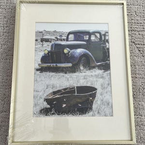 May include: Framed black and white print featuring a vintage truck in a field with a weathered metal trough. The print is encased in a gold-colored frame, adding a touch of rustic charm to the artwork. The image evokes a sense of nostalgia.