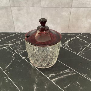 Puede incluir: Un tarro de cristal transparente con un patrón de diamantes tallados en el cuerpo y una tapa de cristal rojo oscuro con un pequeño pomo y una muesca lateral para una cuchara. El recipiente tiene una forma cilíndrica y una base lisa.