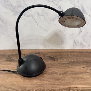 May include: Black desk lamp with a flexible gooseneck arm and a rounded lamp head. The lamp has a circular base and a clear lens. The lamp is on a wooden surface.