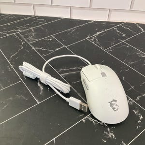 May include: A white wired computer mouse featuring a scroll wheel, two side buttons, and a dragon logo on the palm rest. The mouse is connected to a white USB cable that is neatly coiled and secured with a small white plastic cable tie.