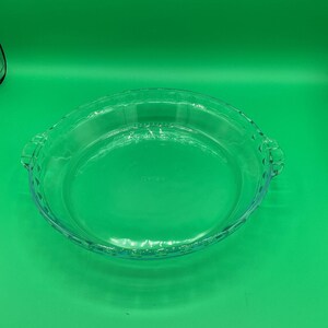 Op de afbeelding: Een heldere glazen taartvorm met een geschulpte rand en twee handvatten. De vorm is rond en heeft het woord "Pyrex" in het glas geëtst. De vorm staat op een groene ondergrond.