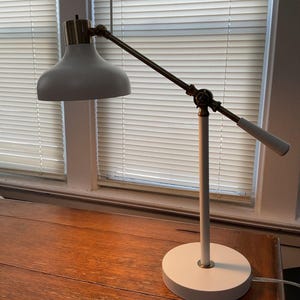 May include: A white and brass desk lamp with an adjustable arm and a round base. The lamp has a white lampshade and a brass-colored joint. The lamp is on a wooden desk near a window with blinds.