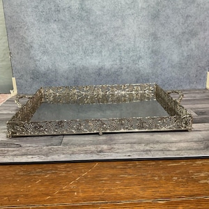 May include: A rectangular silver-toned vanity tray with a mirrored base and ornate grape vine detailing around the edges. The tray has handles on each end and is set on a wooden surface.