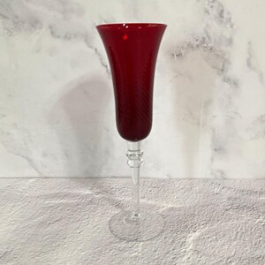 May include: A tall, slender champagne flute featuring a deep red glass bowl with a subtle, diagonal ribbed texture. The stem and circular base are crafted from clear, transparent glass, providing a contrast to the coloured upper section.