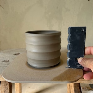 Könnte beinhalten: Eine hellbraune Keramiktasse mit einem strukturierten, gerippten Design. Die Tasse steht auf einem braunen Töpferscheibensockel. Ein schwarzes Plastikwerkzeug mit einem gewellten Rand ist im Hintergrund zu sehen.