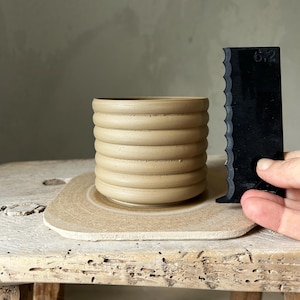 Könnte beinhalten: Ein hellbrauner Keramiktopf mit geripptem Design steht auf einer Töpferscheibe. Neben dem Topf liegt ein schwarzes Plastikwerkzeug mit gezacktem Rand.