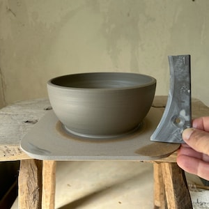 Könnte beinhalten: Eine graue Keramikschüssel steht auf einem grauen Töpferscheibensockel. Ein graues Metallwerkzeug liegt neben der Schüssel.