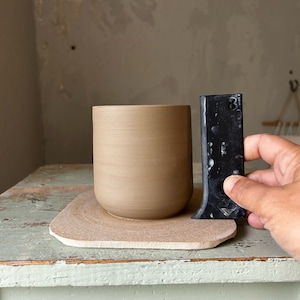 Könnte beinhalten: Eine beige Keramiktasse steht auf einem hellbraunen, rechteckigen Untersetzer. Ein schwarzes Werkzeug wird daneben gehalten. Die Tasse hat eine zylindrische Form mit strukturierter Oberfläche. Der Untersetzer hat eine abgerundete Kante.