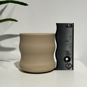 Könnte beinhalten: Eine hellbraune Keramiktasse mit welligem Design. Die Tasse ist 5,3 Zoll hoch und steht neben einem schwarzen Lineal.