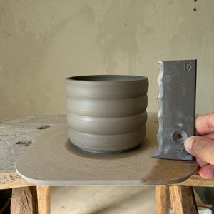 Könnte beinhalten: Eine hellbraune Keramiktasse mit geripptem Design steht auf einer Töpferscheibe. Ein Metallwerkzeug mit gezacktem Rand und der Zahl "5" darauf befindet sich neben der Tasse.