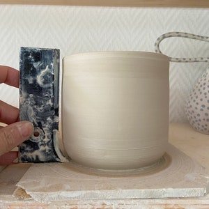 Puede incluir: Una taza de cer&aacute;mica beige claro con base redondeada sobre una estanter&iacute;a de madera. Una herramienta gris oscuro se sostiene junto a la taza. La taza es cil&iacute;ndrica con una superficie lisa y sin esmaltar.