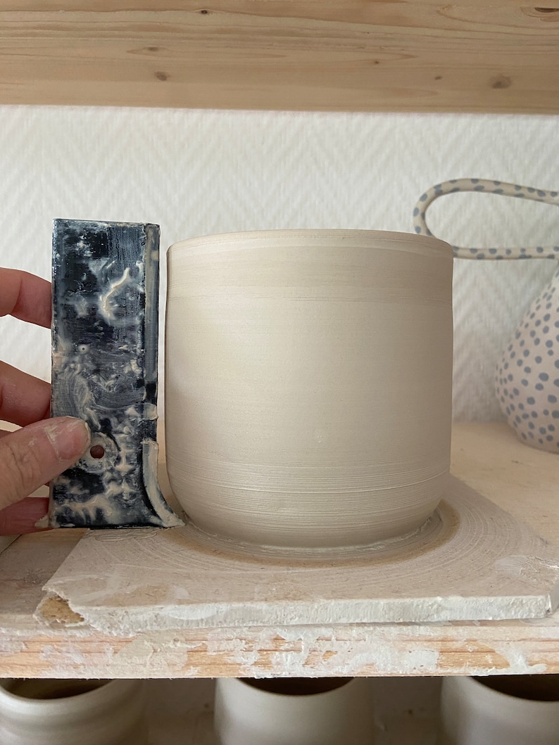 Puede incluir: Una taza de cer&aacute;mica beige claro con base redondeada sobre una estanter&iacute;a de madera. Una herramienta gris oscuro se sostiene junto a la taza. La taza es cil&iacute;ndrica con una superficie lisa y sin esmaltar.