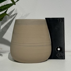 Könnte beinhalten: Eine hellbeige Keramiktasse mit einem schwarzen Plastikhalter. Der Halter hat die Nummer "13" aufgedruckt.