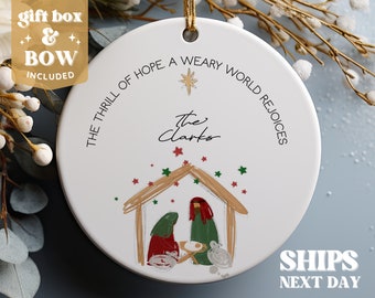 May include: White ceramic Christmas bauble featuring a nativity scene illustration. The bauble has the text "The Thrill of Hope. A Weary World Rejoices" and "The Clarks" written on it.