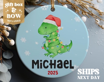 May include: A blue and white Christmas ornament featuring a green dinosaur wearing a red Santa hat and Christmas lights. The dinosaur's name, "Michael", and the year "2025" are printed on the ornament.