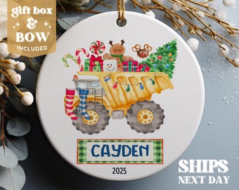 May include: A yellow toy dumper truck with a Christmas tree, presents, and gingerbread people in the back. The truck has a red and blue stocking on each side. The truck is on a white background with the name "Cayden" and the year "2025" printed below.