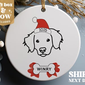 Pu&ograve; includere: Addobbo natalizio in ceramica bianca con un disegno a linea nera di un cane che indossa un cappello di Babbo Natale rosso con l'anno 2025. Il nome del cane, "WINRY", &egrave; scritto su un osso rosso e bianco.