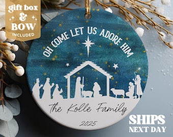 May include: A round Christmas ornament with a blue and white design. The ornament features a nativity scene silhouette and the text "OH COME LET US ADORE HIM" and "The Kolle Family 2025". A gold ribbon is attached for hanging.